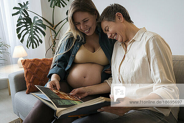 Pregnant daughter sharing photo album with mother sitting on sofa at home