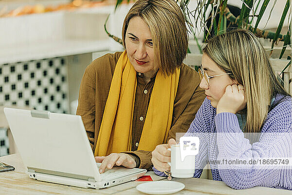 Mutter teilt Laptop mit Tochter und hält Kaffeetasse im Café