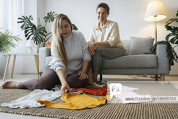 Mother and pregnant daughter arranging baby clothes on floor at home