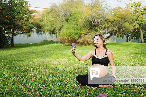 Happy pregnant woman taking selfie sitting cross-legged on grass
