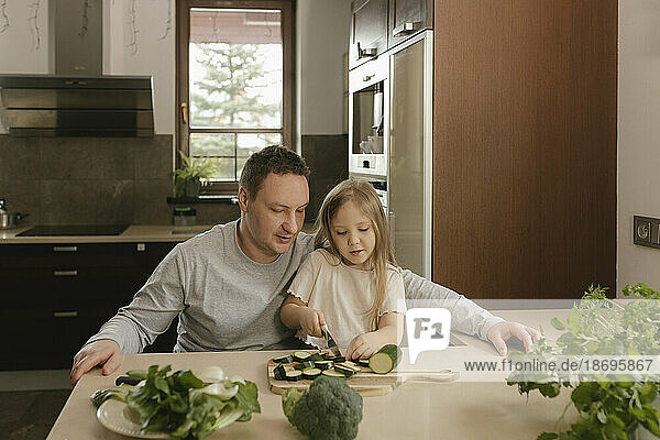 Vater sitzt mit Tochter und schneidet Zucchini am Küchentisch