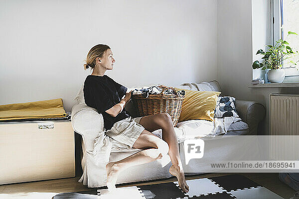 Nachdenkliche Frau mit Wäschekorb entspannt sich zu Hause auf dem Sofa
