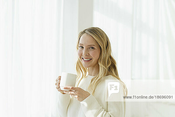 Smiling woman holding coffee cup by translucent curtain