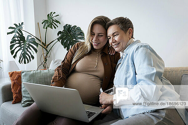 Lächelnde Frau beim Online-Shopping mit schwangerer Tochter über Laptop zu Hause