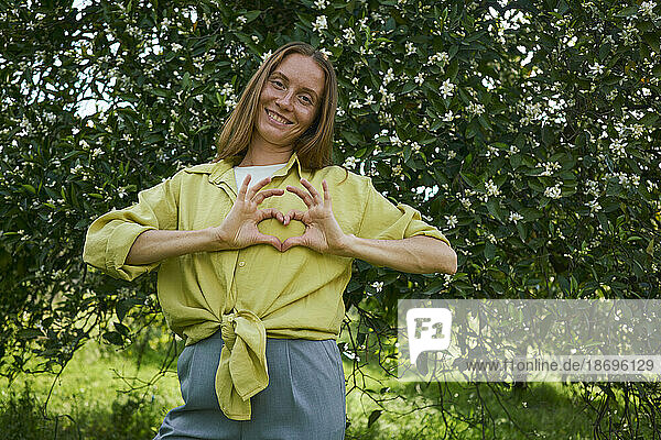 Woman making heart gesture standing in front of tree at garden