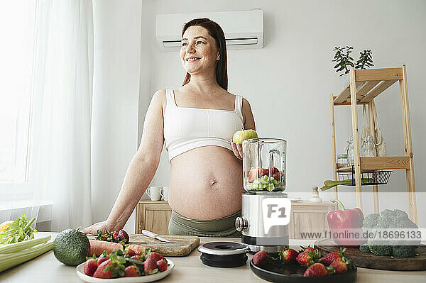 Smiling pregnant woman preparing fruit smoothie in kitchen at home