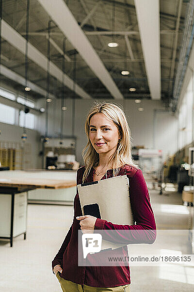 Smiling businesswoman with hand in pocket holding file in factory