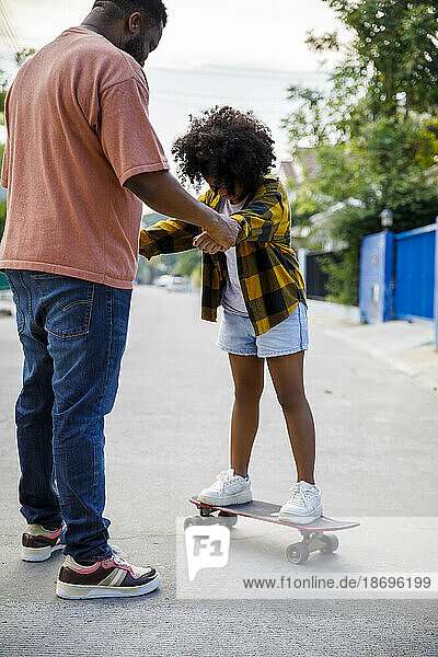 Father assisting daughter standing on skateboard at footpath