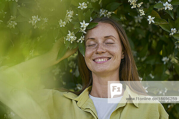 Lächelnde Frau mit geschlossenen Augen und orangefarbener Blüte im Garten