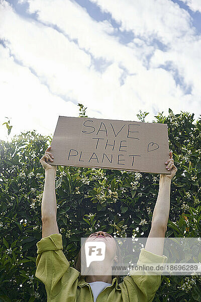 Frau hält einen ausgeschnittenen Karton mit dem Text „Save The Planet“ neben einem Baum im Garten