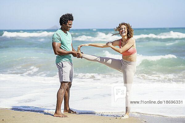 Man assisting girfriend stretching and standing on one leg at seashore