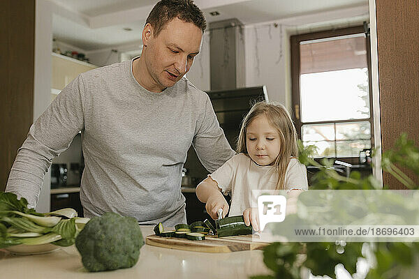 Father teaching daughter to cut zucchini in kitchen at home