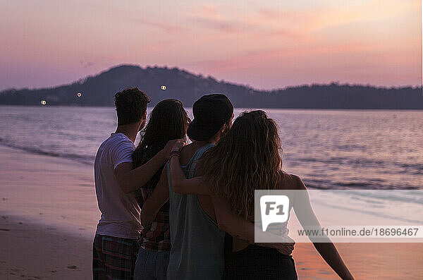 Friends with arms around standing at sunset beach