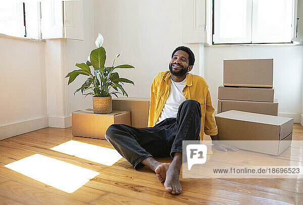 Smiling man sitting by cardboard boxes on floor in new apartment