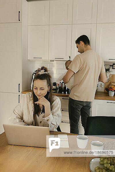 Woman working on laptop in kitchen with family in background at home