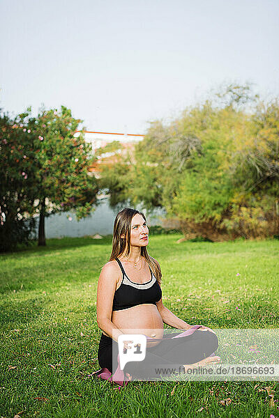 Nachdenkliche schwangere Frau sitzt mit gekreuzten Beinen auf dem Rasen