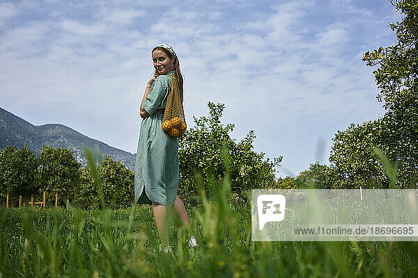 Frau mit Orangen im Netzbeutel steht im Obstgarten