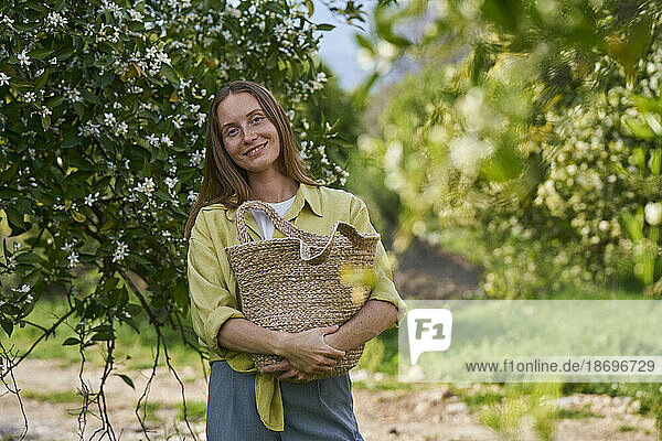 Smiling woman holding straw bag standing in orchard