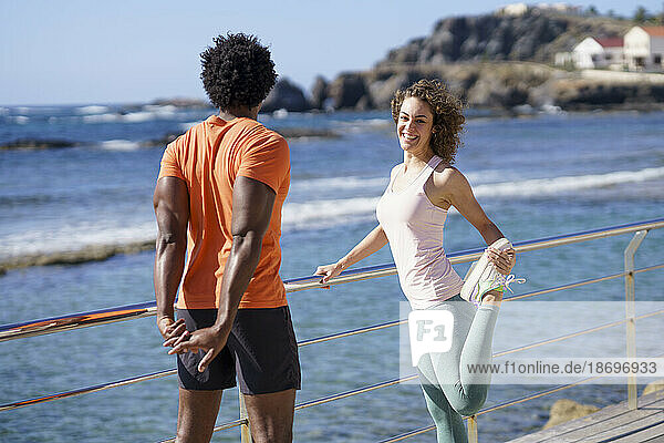 Smiling woman looking at boyfriend doing stretching exercise on pier in coastal area