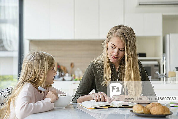 Mother reading book sitting with daughter in kitchen at home