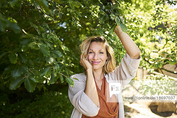 Smiling woman with hand in hair standing under tree