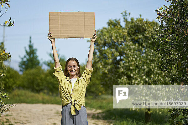 Lächelnde Frau hält ausgeschnittenen Karton mit erhobenen Armen im Garten