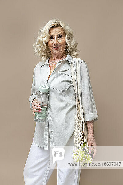 Ältere Geschäftsfrau mit Netzbeutel und Wasserflasche vor beigem Hintergrund