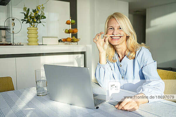 Freelancer woman playing with pen by laptop at desk in home office
