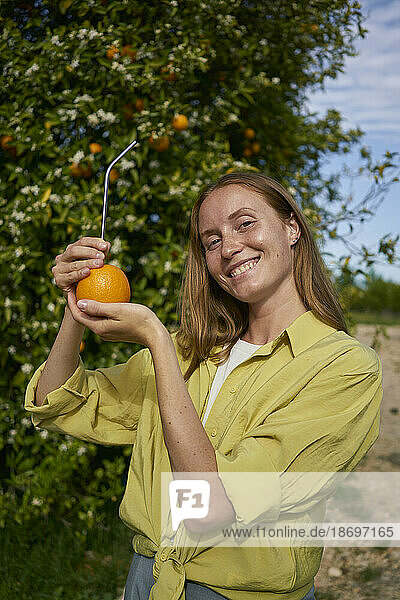 Happy woman showing orange with reusable metal straw in orchard