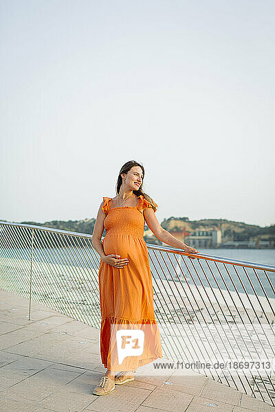 Smiling pregnant woman with hand on stomach standing by railing