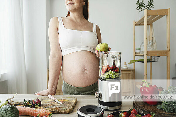 Pregnant woman leaning on table with blender in kitchen at home