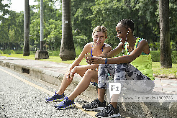 Fröhliche Freunde  die ihr Smartphone teilen und auf dem Bürgersteig im Park sitzen