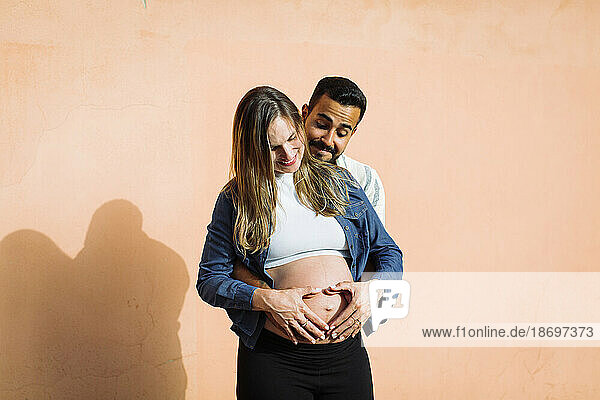 Smiling man making heart shape of pregnant woman's belly in front of wall