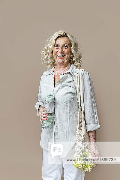 Smiling senior businesswoman with holding water bottle and mesh bag with fruits against beige background
