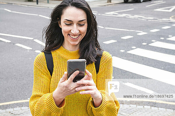 Smiling female tourist in yellow sweater using smartphone near city street