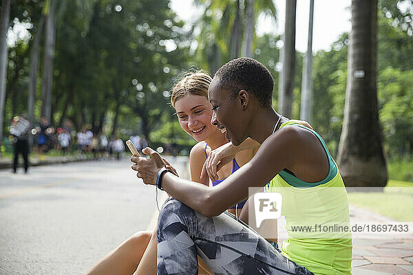 Happy woman using smart phone sitting with friend on sidewalk at park