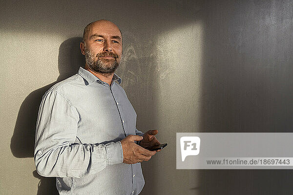 Mature businessman holding mobile phone at a wall