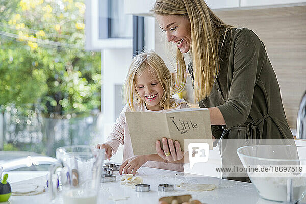 Glückliche Frau liest Rezeptbuch von Tochter  die zu Hause in der Küche Essen zubereitet