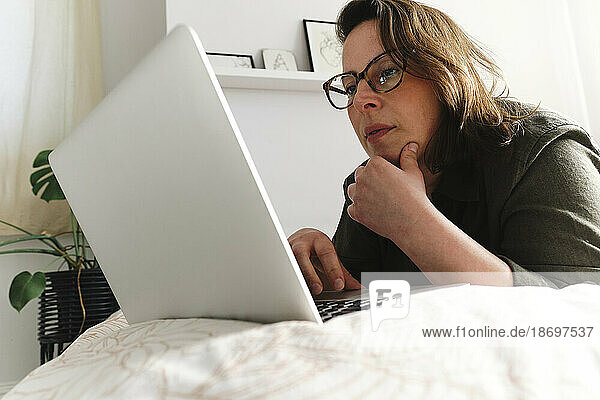 Freelancer with hand on chin using laptop on bed at home