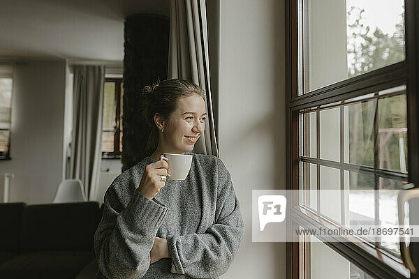Smiling woman with coffee cup looking out of window at home