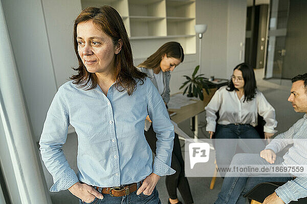 Thoughtful businesswoman looking out of window in office with colleagues in background