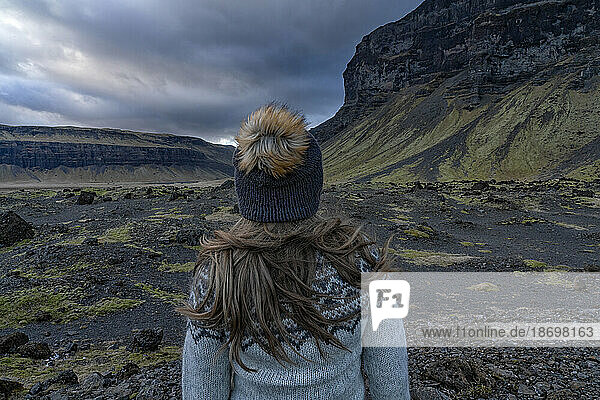 Close-up view of a woman with blond hair  taken from behind  wearing a woolen hat and an Icelandic sweater while standing out in the landscape admiring the natural beauty of the mountains and stormy sky; South Iceland  Iceland