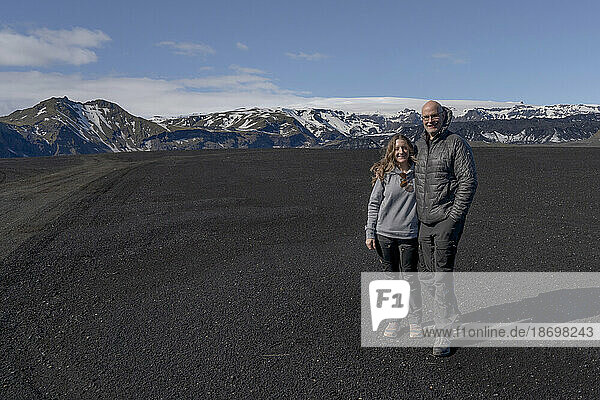 Close-up view of a smiling couple posing for the camera  standing in front of a massive landscape of black sand with a mountain ridge and a blue sky in the background; Iceland