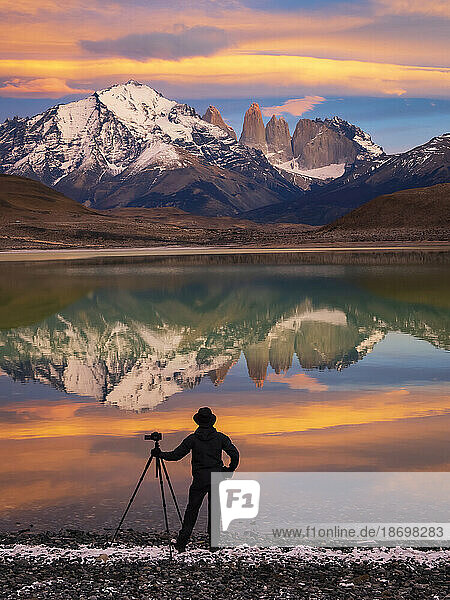View taken from behind of a silhouette of a photographer with a camera tripod standing at the water's edge at Lago Azul at sunrise tracking pumas; Torres del Paine National Park  Patagonia  Chile