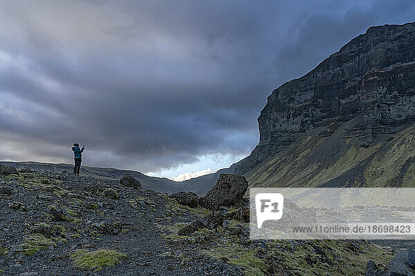 Woman standing out on the volcanic  rocky landscape admiring the natural beauty and taking a picture of the mountains and dramatic storm clouds forming overhead; South Iceland  Iceland
