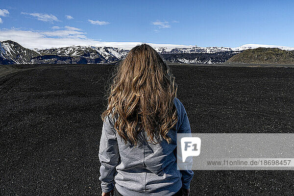 Close-up view of a woman with blond hair  taken from behind  standing in front of a massive landscape of black sand with a mountain ridge and a blue sky in the distance; Iceland