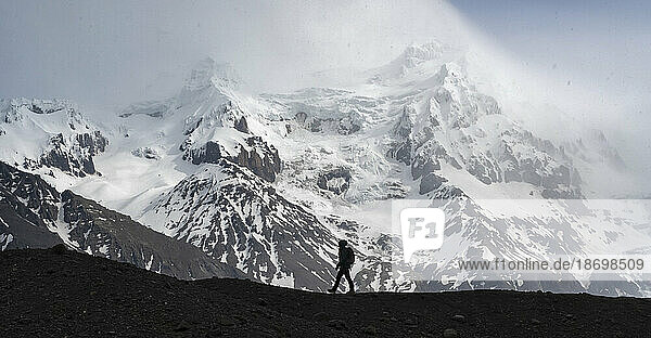 Silhouette of a person walking along a glacial moraine in front of a large snow-capped mountain on the South Coast of Iceland; South Iceland  Iceland