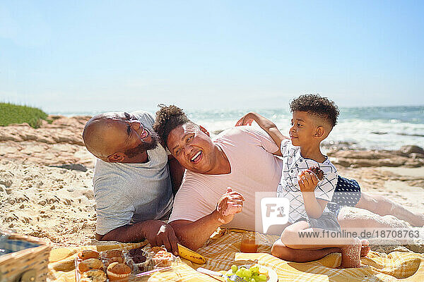 Happy gay male couple and son laughing and eating on sunny beach