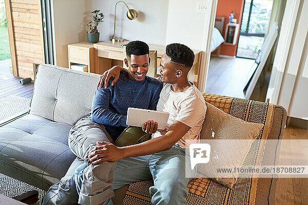 Happy young gay male couple hugging and using digital tablet on sofa