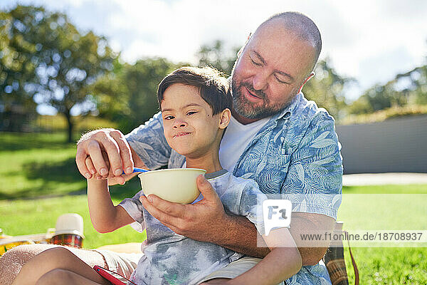 Portrait father feeding cute son with Down Syndrome in summer park